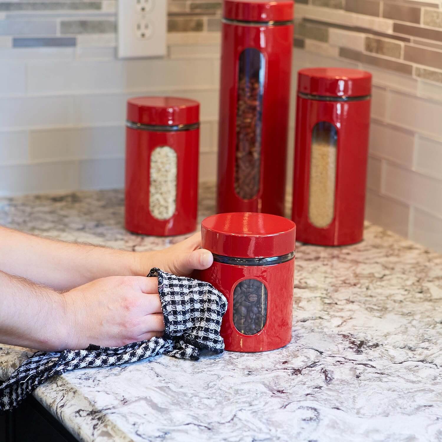 Person cleaning a red Anchor Hocking canister set on a kitchen countertop with a checkered cloth.
