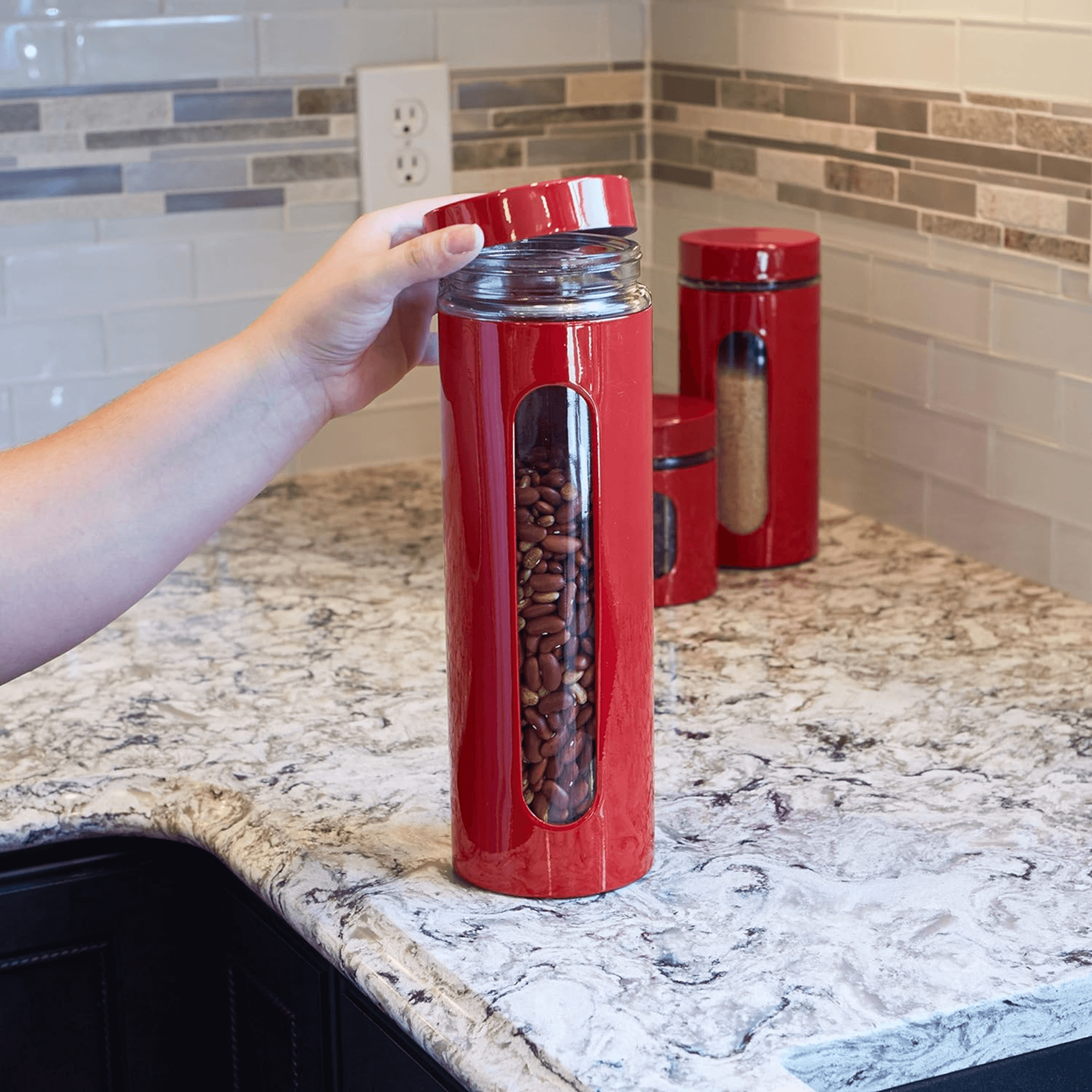 Hand holding a red glass canister filled with beans, showcasing the stylish Anchor Hocking Canister Set on a kitchen countertop.