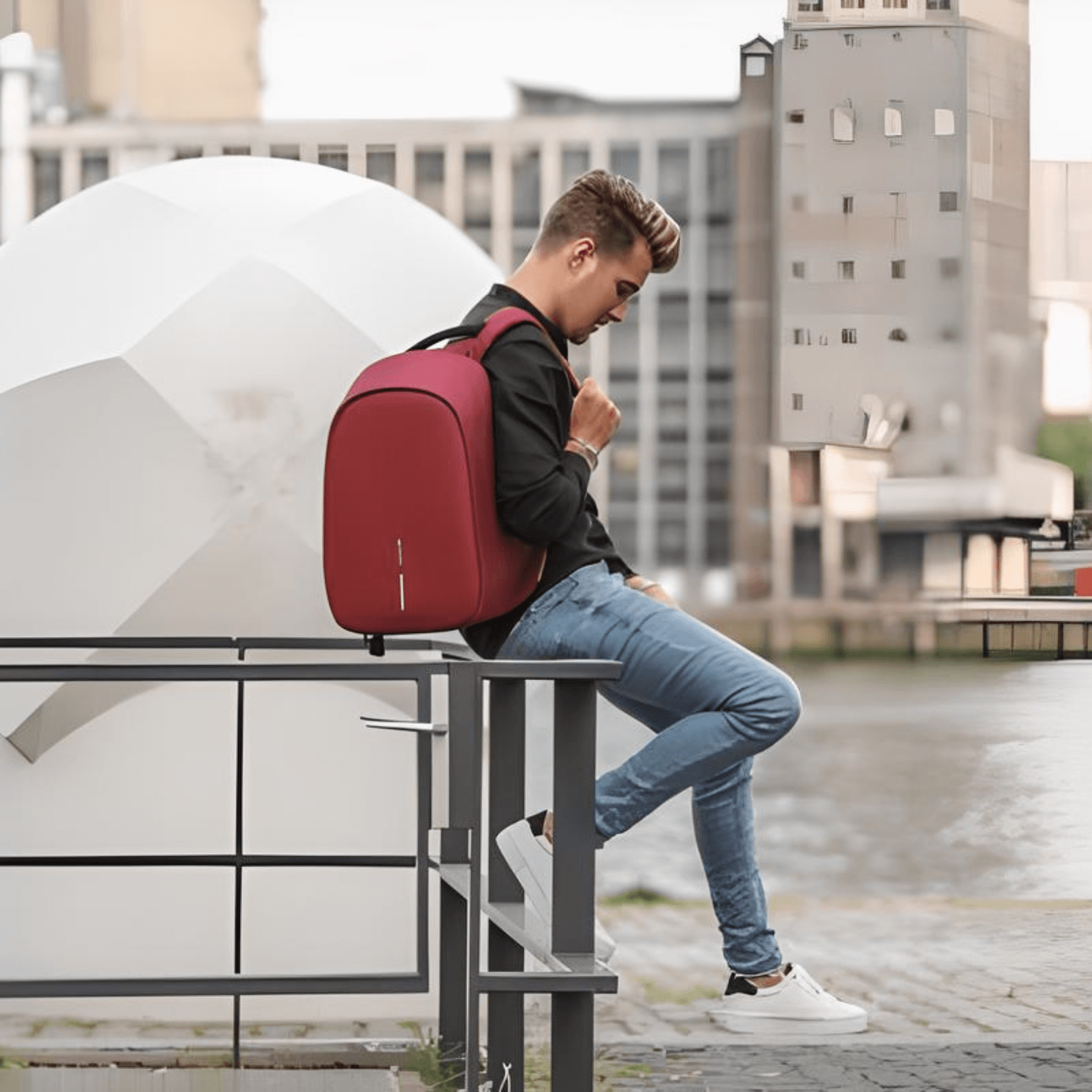 A young man sitting on a railing with the Bobby Hero Regular Anti-Theft Backpack in Cherry Red, showcasing modern design and style.