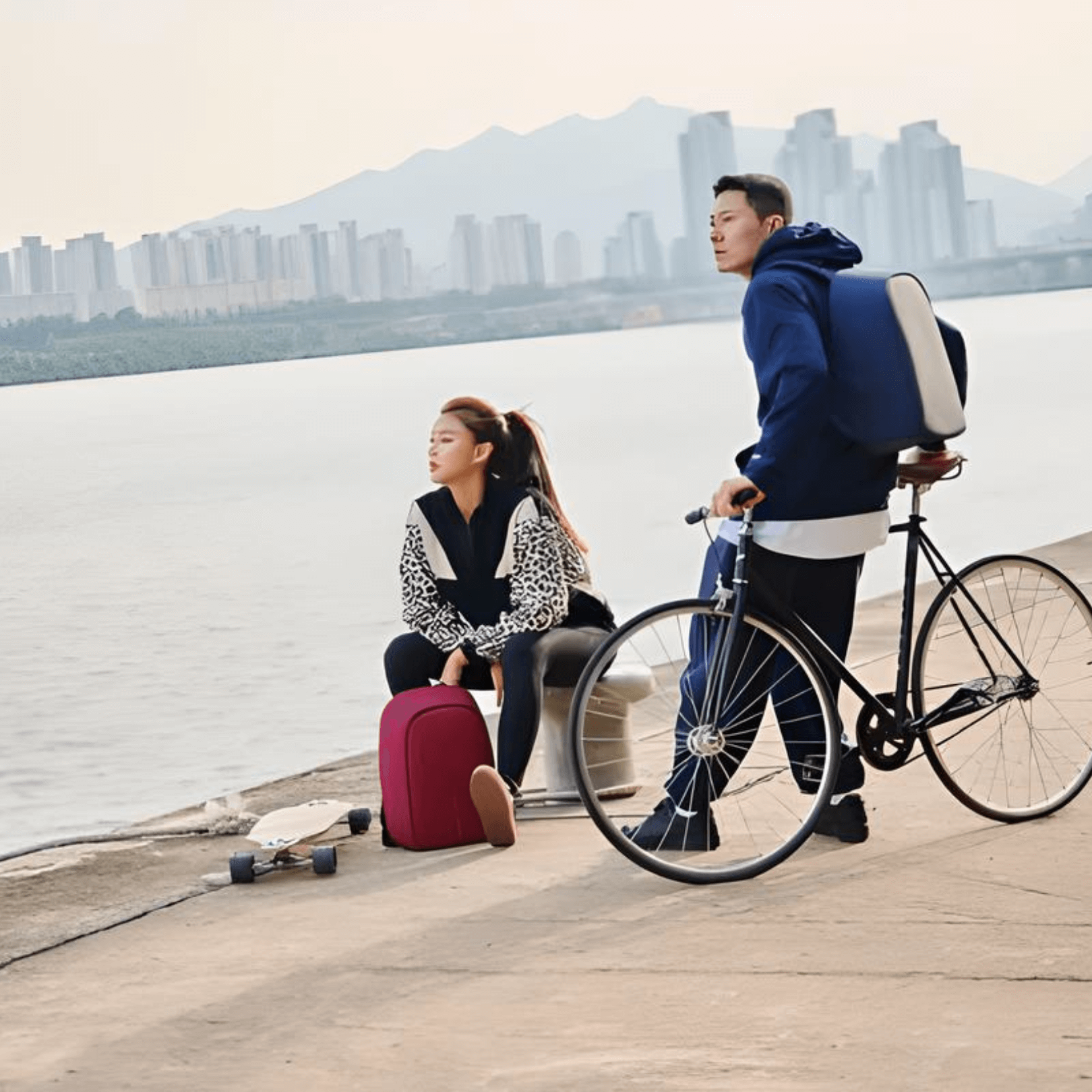 A woman sitting near a river with a red backpack and a man with a bicycle, showcasing an outdoor lifestyle.