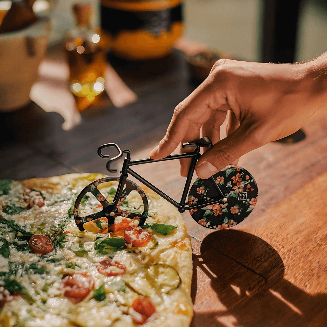 Hand using a floral bicycle-shaped pizza cutter on a vibrant pizza, showcasing the Fixie Pizza Cutter design.