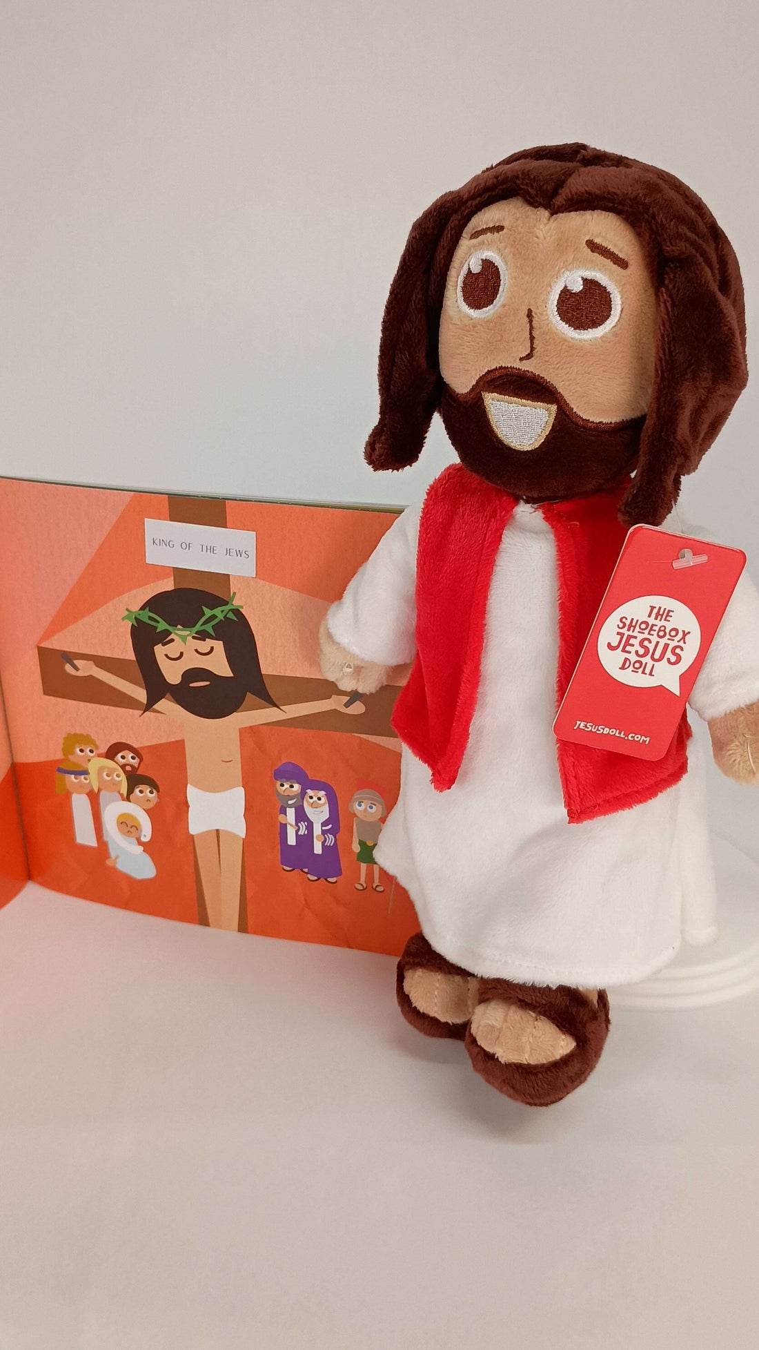 Cuddly Jesus doll with red scarf next to Bible storybook for kids, promoting faith and storytelling.