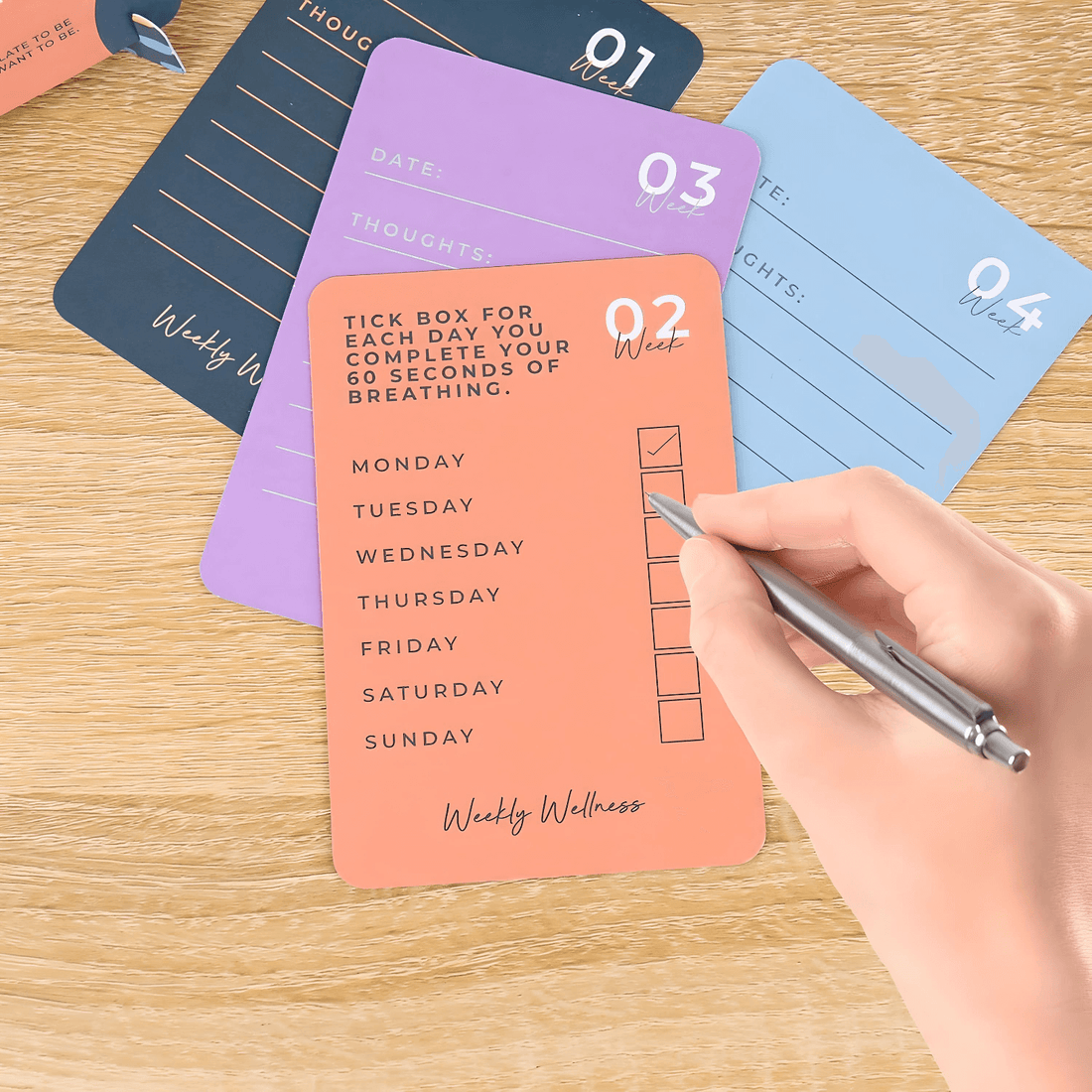 Hand holding a pen marking off days on Mindfulness Weekly Wellness Cards for self-care and mental clarity challenges.