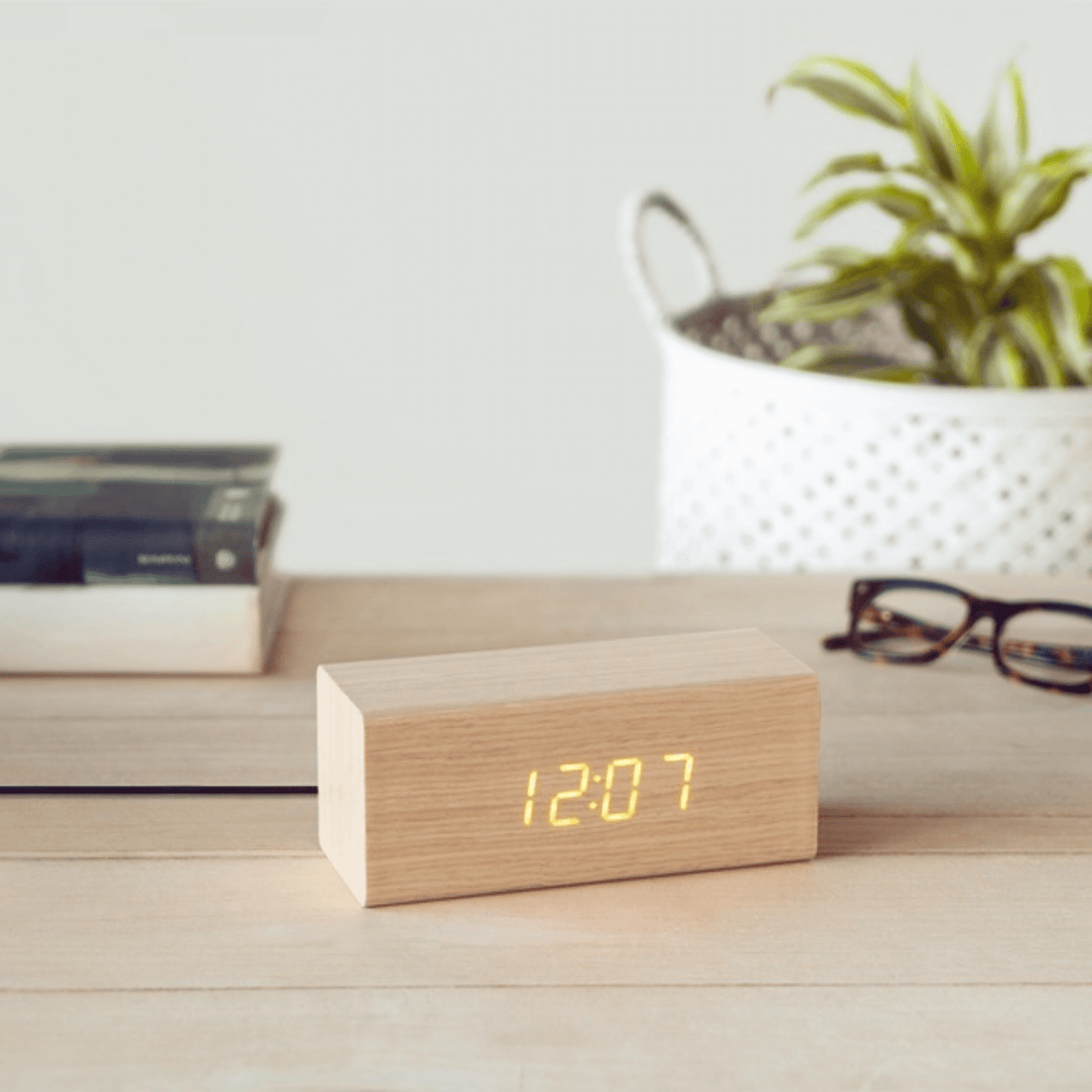 Minimalist wooden LED alarm clock in cube design displaying time on a wood surface with plant and books in the background.