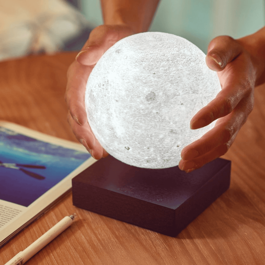 Person holding a glowing Smart Moon Lamp on a wooden table with a magazine and pen nearby.