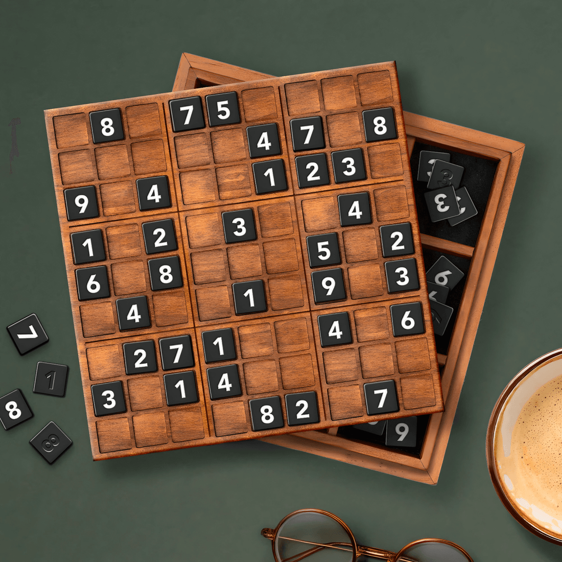 Sudoku Deluxe XL wooden board game with black tiles, set on a green background next to a cup of coffee and glasses.
