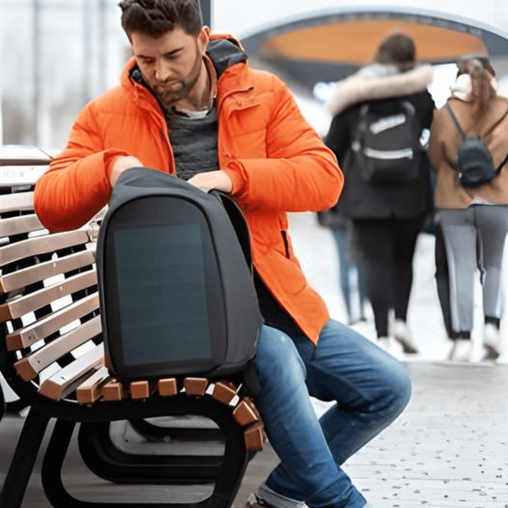 Man in an orange jacket sitting on a bench, using the XD Design Bobby Tech Anti-Theft Backpack.